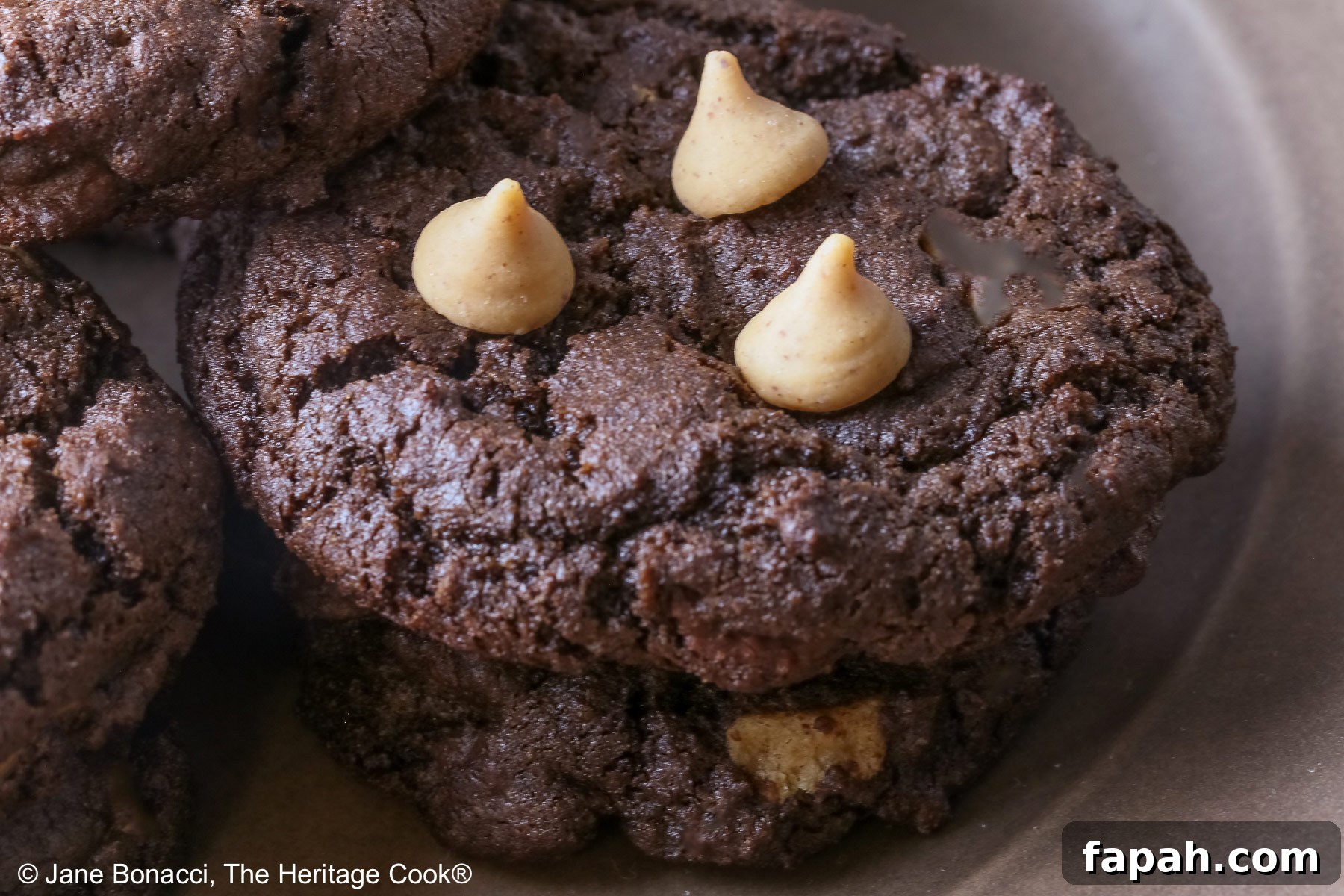 Gluten-Free Double Chocolate Peanut Butter Cookies Recipe 7 Four stacks of black Double Chocolate Peanut Butter Cookies with light peanut butter chips on top on a deep brown ceramic plate sitting on a bright pink cloth © 2025 Jane Bonacci, The Heritage Cook.