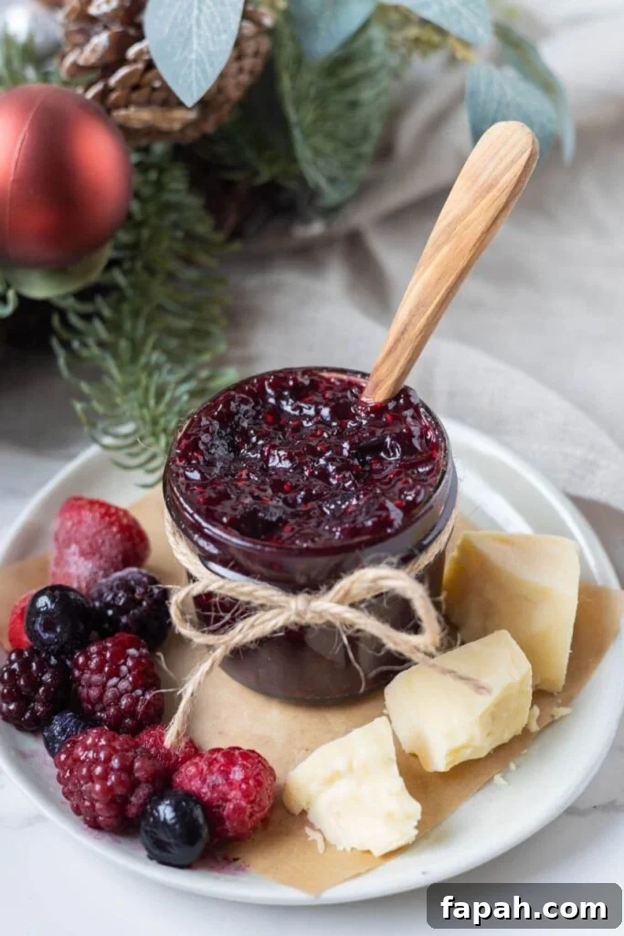 A small jar of mulled berry jam tied with a twine bow. The jar of jam is sitting on a plate alongside a selection of mixed berries and crumbly cheddar cheese. There is a Christmas wreath in the background of the jar.