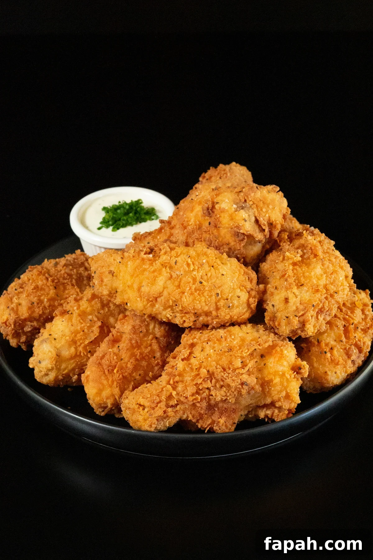 A small black plates has about a dozen breaded and fried chicken wings stacked on top of each other. A small side of ranch sauce topped with chives is out of focus in the background.
