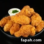 A small black plates has about a dozen breaded and fried chicken wings stacked on top of each other. A small side of ranch sauce topped with chives is out of focus in the background.