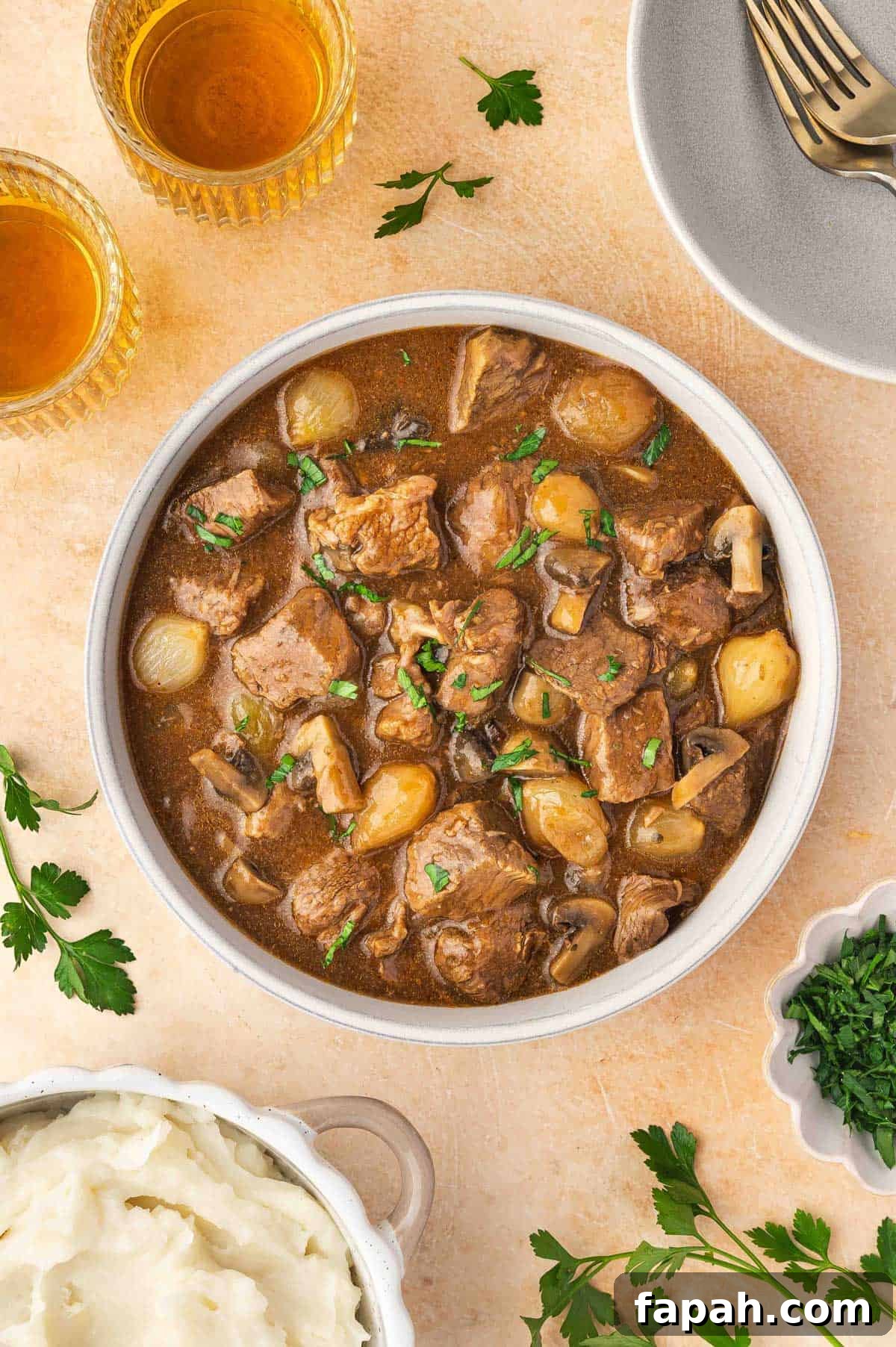 Overhead view of a bowl of slow cooker beef tips and gravy surrounded by a bowl of mashed potatoes, two drinks, and fresh herbs.