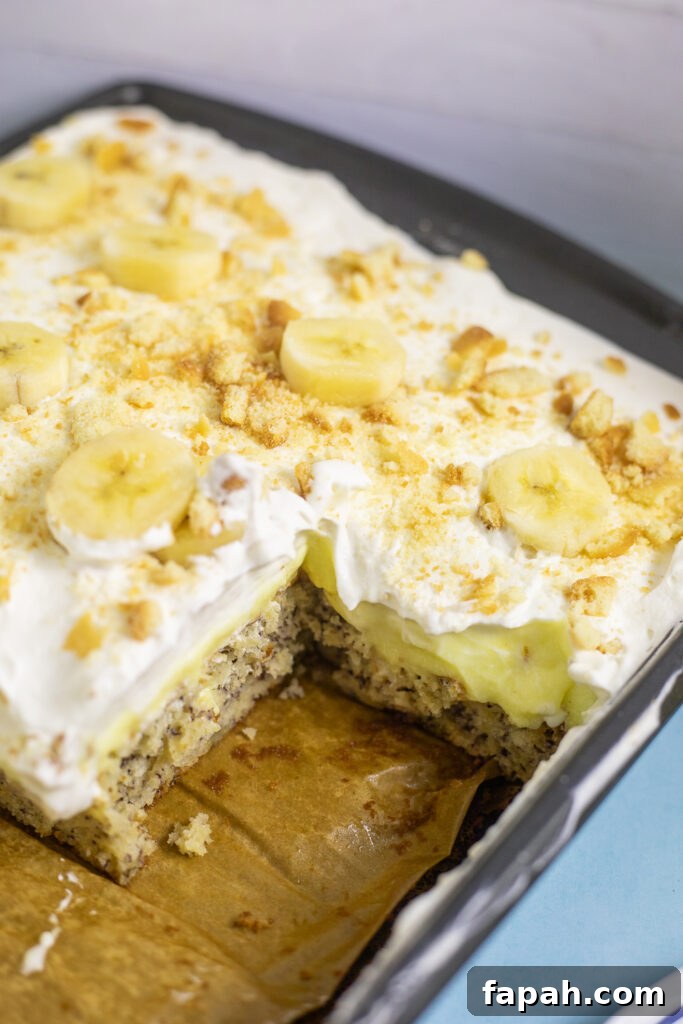 Banana pudding cake in a sheet cake pan with a piece missing showing the inside of the cake with vanilla wafer crumbs and fresh banana slices on top.