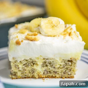 A piece of banana pudding poke cake topped with whipped cream, vanilla wafer crumbs, and fresh banana slices showing the pudding in the center of the cake, on a small white plate with bananas and the remaining cake behind.