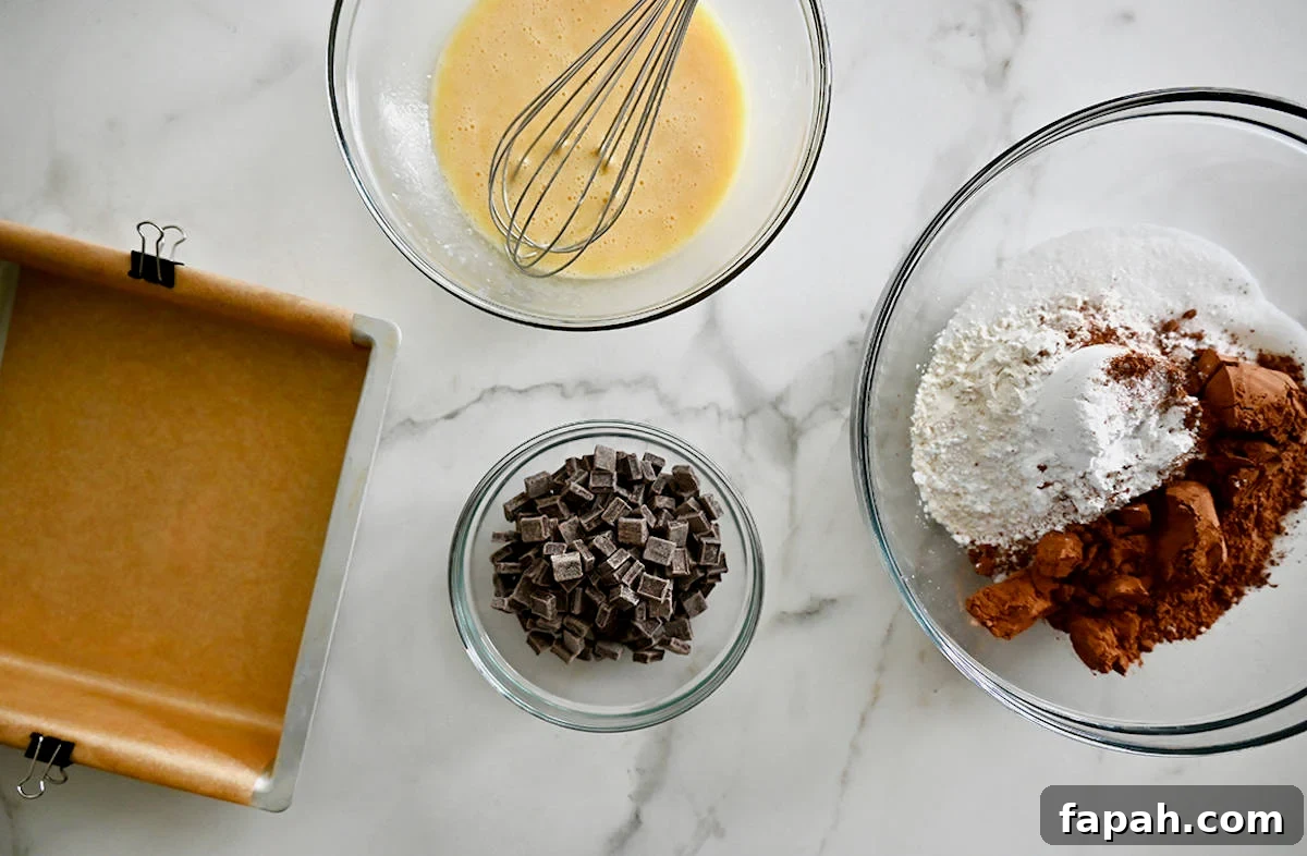 Fudgy Cocoa Brownies Recipe: Rich, Chewy Homemade Treat 5 A square baking dish lined with parchment paper next to three glass bowls containing chocolate chunks, flour and cocoa powder, and a whisked egg.