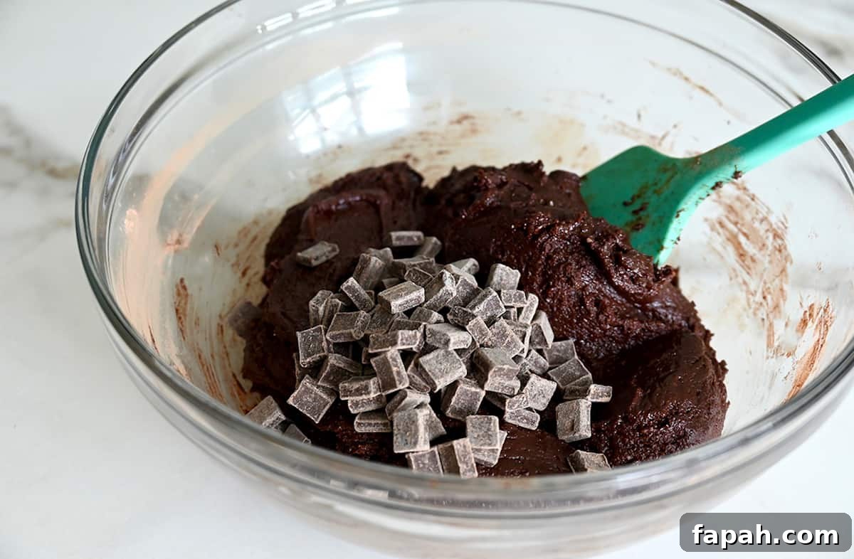 Fudgy Cocoa Brownies Recipe: Rich, Chewy Homemade Treat 6 A teal spatula folds chocolate chunks into brownie batter in a glass bowl.