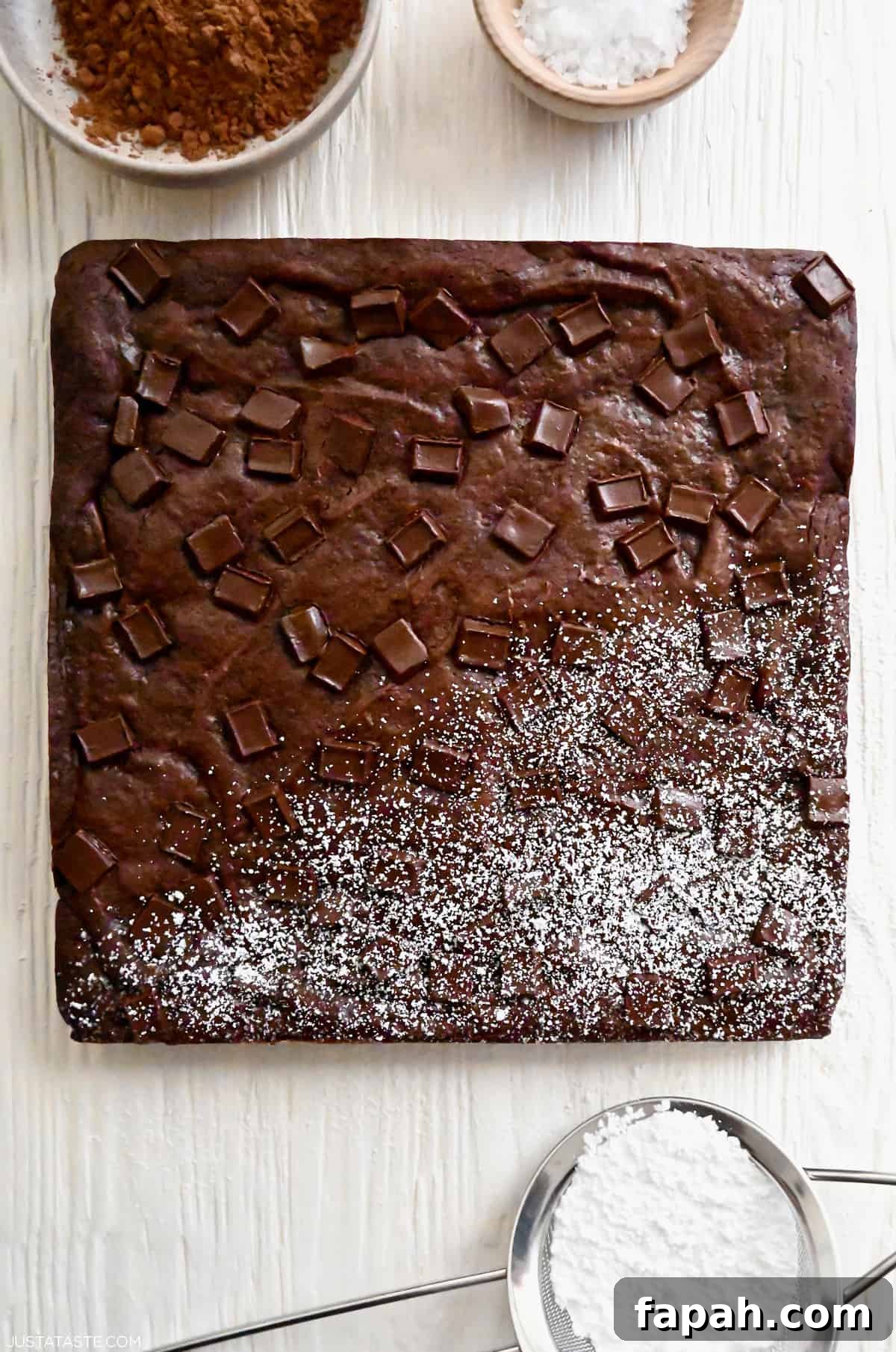 Fudgy Cocoa Brownies Recipe: Rich, Chewy Homemade Treat 7 An uncut square brownie with chocolate chunks that's half dusted with powdered sugar.