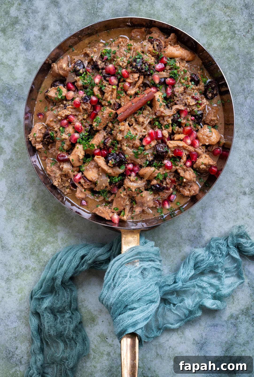 Savory Persian Chicken Stew Recipe (Khoresh-e Morgh) 3 A pan with a golden handle contains a colorful stew of chicken, garnished with pomegranate seeds, herbs, and a cinnamon stick. The pan rests on a textured, green surface, with a teal cloth wrapped around the handle.