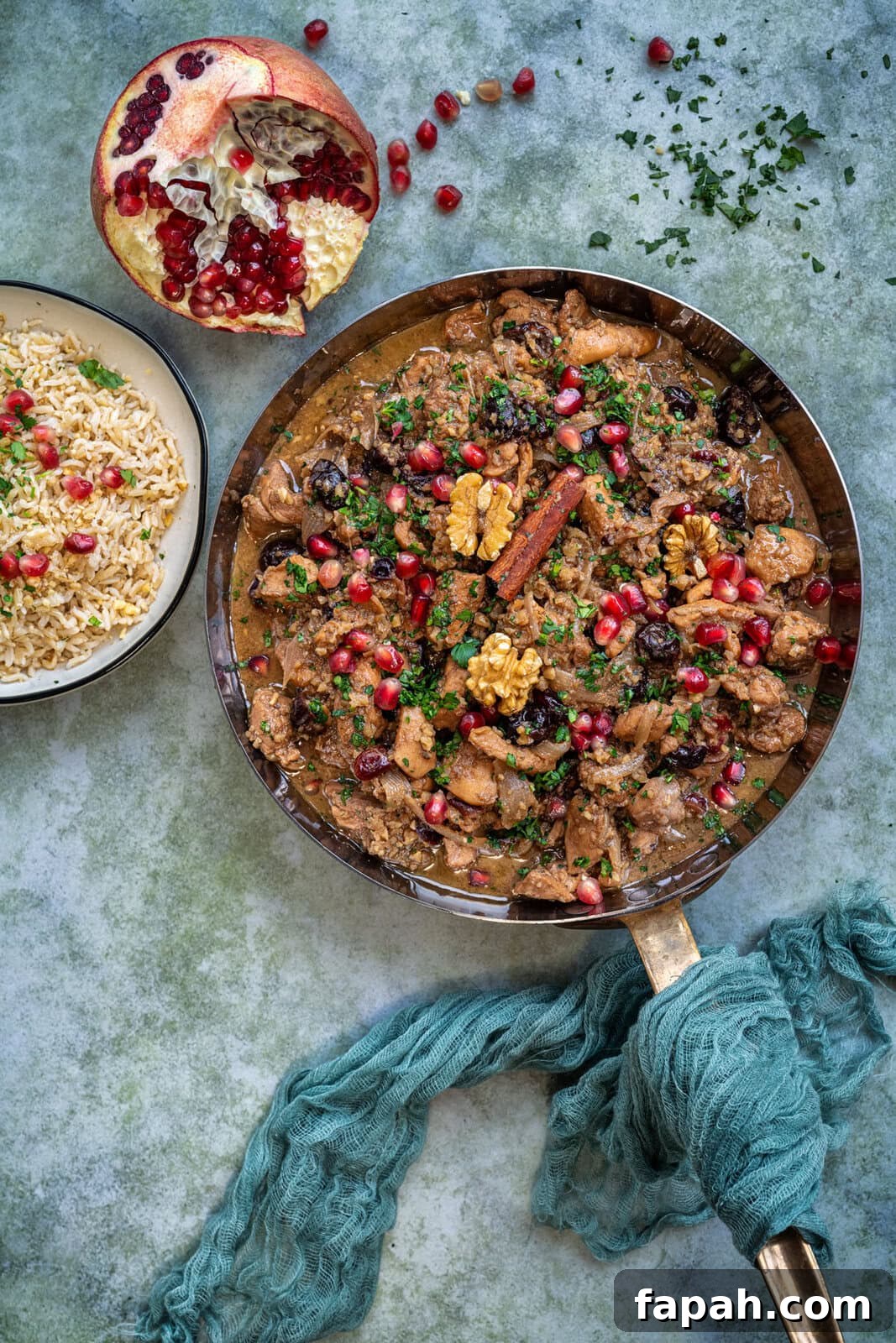 Savory Persian Chicken Stew Recipe (Khoresh-e Morgh) 9 A skillet with Persian stew garnished with walnuts and pomegranate seeds. A bowl of rice topped with herbs and seeds is beside it. A halved pomegranate is also visible. A teal cloth is wrapped around the skillets handle on a textured surface.