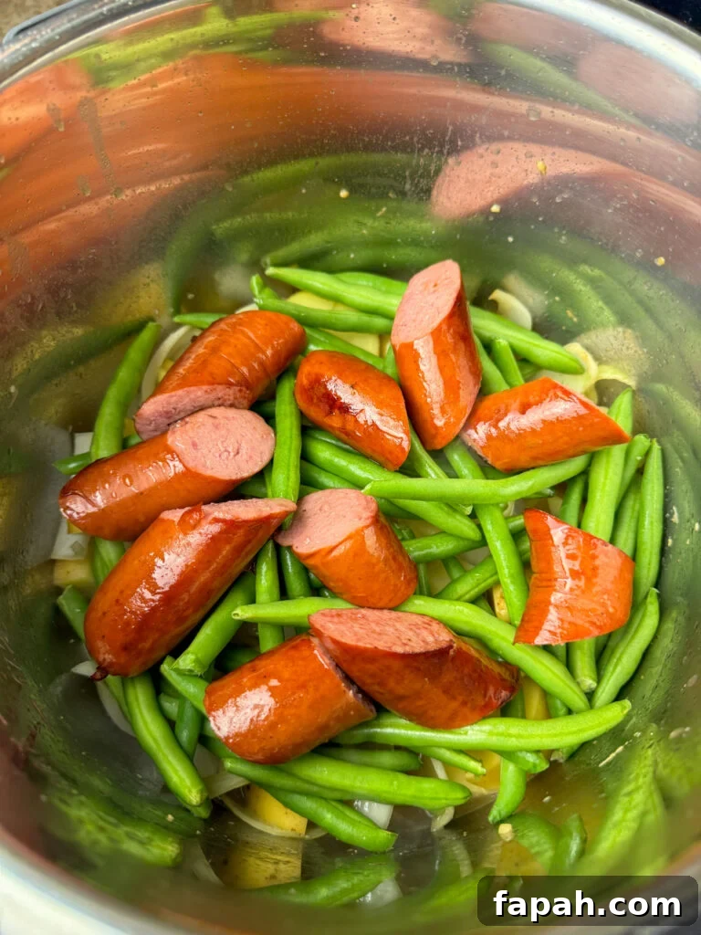 Browned sausage laying on top of fresh green beans and raw potatoes in a silver pressure cooker.