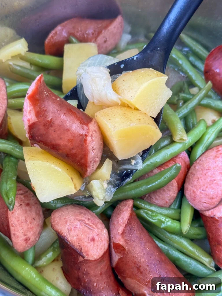 Black serving spoon lifting a warm chunk of sausage and several chunks of potatoes with slices of onion and pieces of garlic above the pot full and green beans underneath.