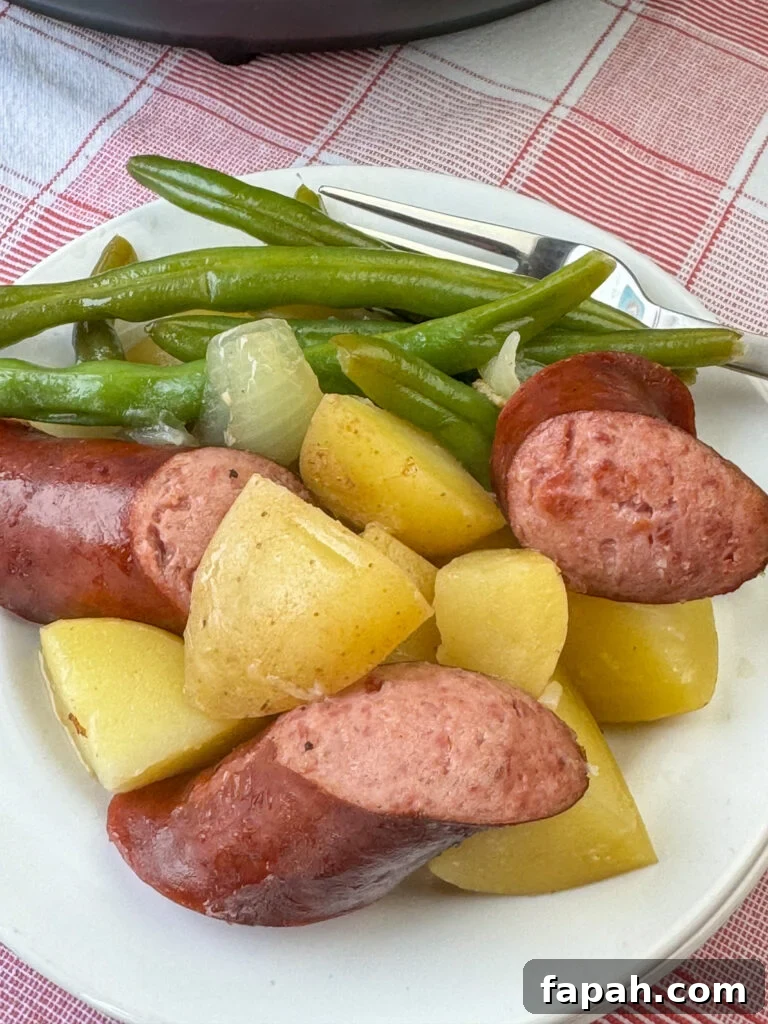 Tender potatoes nestled among chunks of smoked sausage with onions and green beans on the back of the plate next to the fork resting on a red and white plaid napkin.