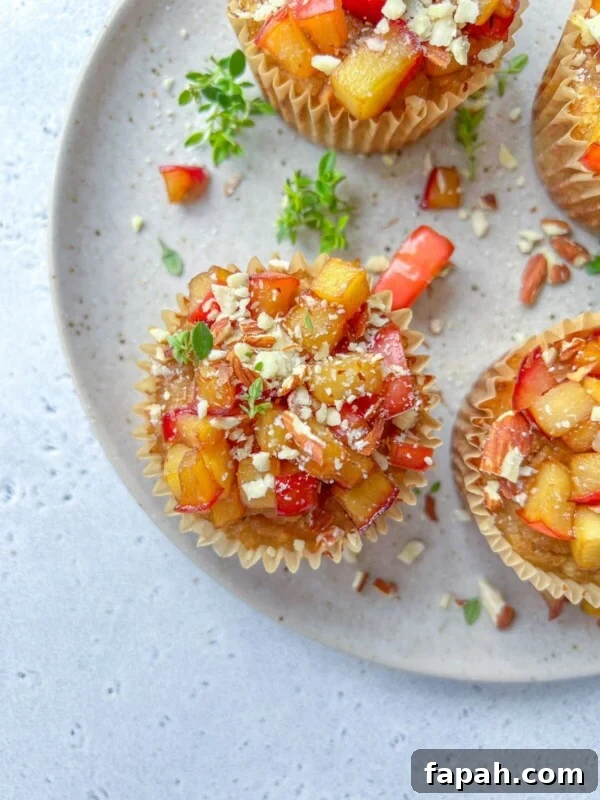 Overhead view of Almond Flour Blueberry Muffins topped with diced fresh peaches and crushed nuts, arranged on a speckled ceramic plate with a few green herb sprigs scattered around.