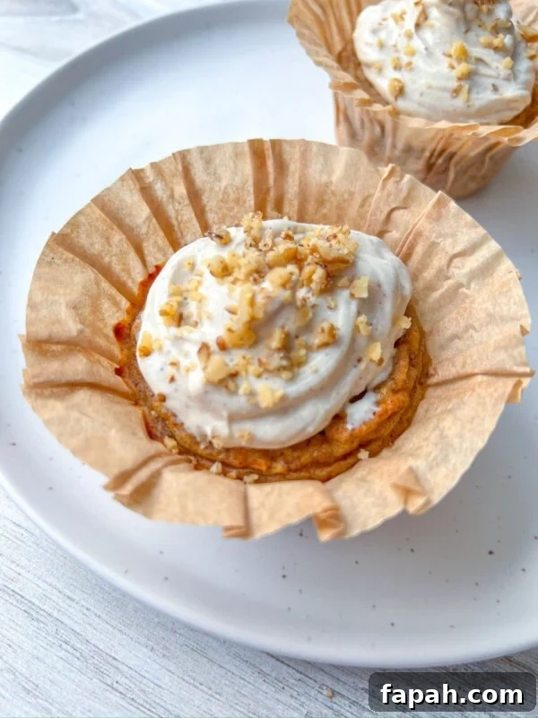 A Healthy Carrot Cake Muffin with white frosting and chopped nuts sits in a brown paper liner on a white plate, with another similar muffin blurred in the background.