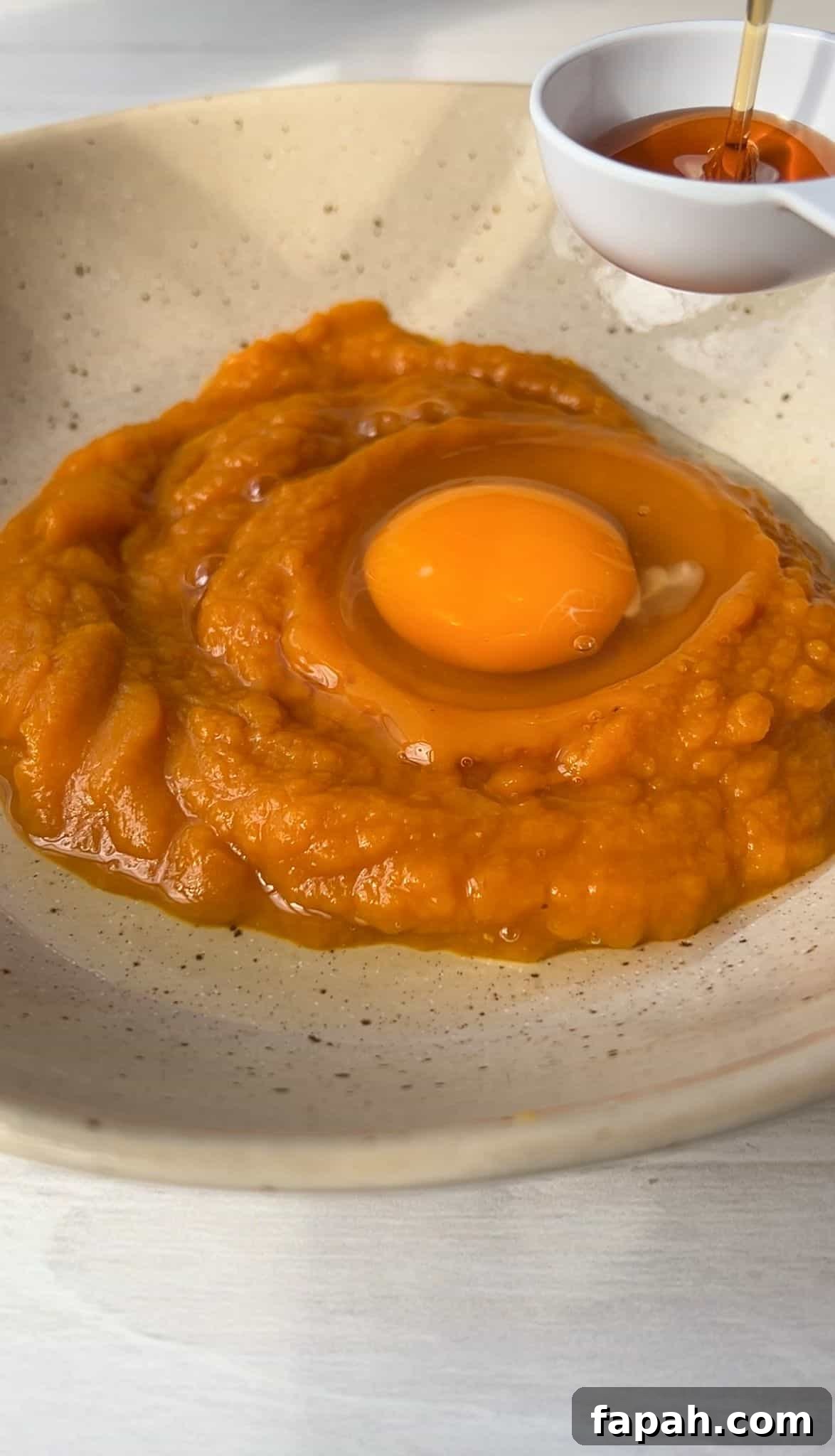 A bowl containing pumpkin puree with a raw egg on top. Honey is being drizzled from a small white bowl above the mixture. The bowl is placed on a light-colored surface.