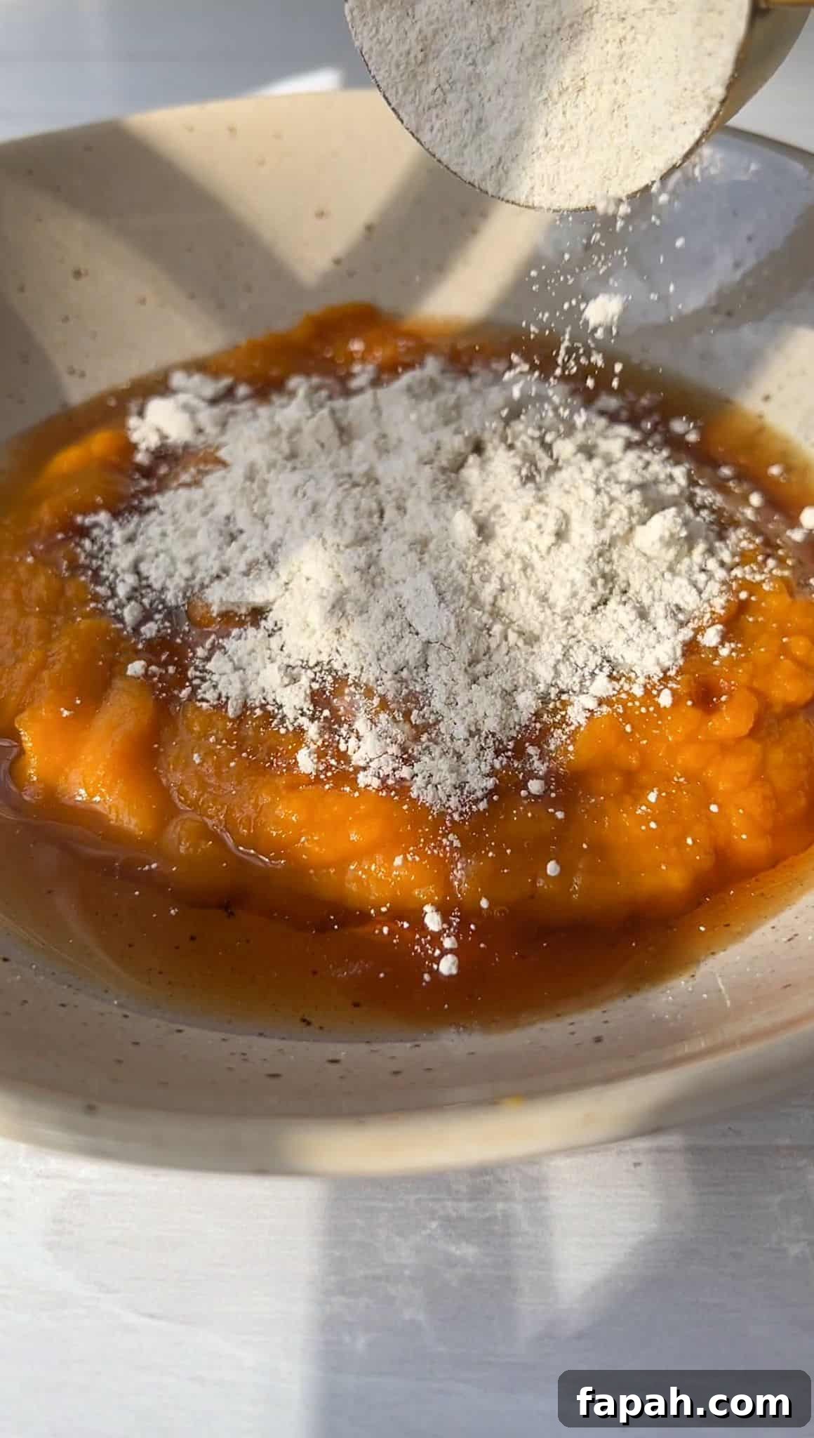 A bowl contains orange pumpkin puree topped with a spoonful of white flour. Warm sunlight casts soft shadows, highlighting the rustic texture of the brown-speckled ceramic bowl and the smooth surface of the puree.
