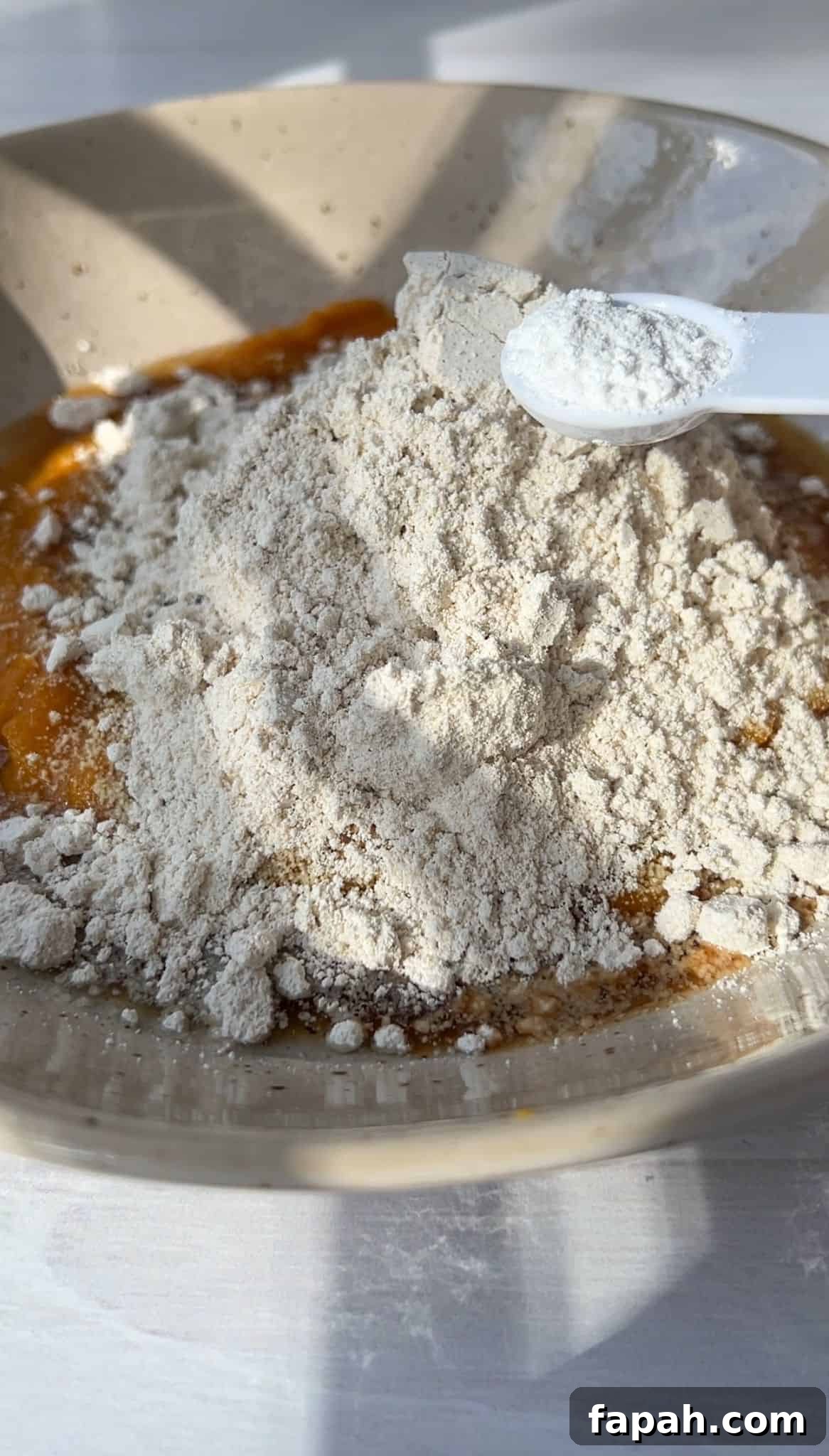 A bowl filled with a mixture of flour on top of a liquid, possibly dough or batter, with a measuring spoon resting on the flour. Sunlight casts shadows, highlighting the powdery texture of the flour.