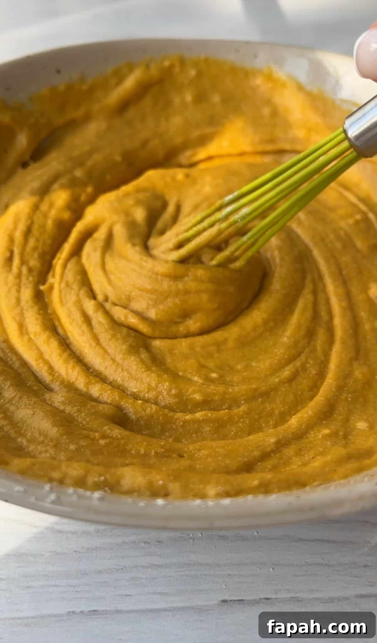 A close-up of a creamy, orange-hued mixture being whisked in a beige bowl. The texture appears smooth and thick. The bowl is placed on a light wooden surface.