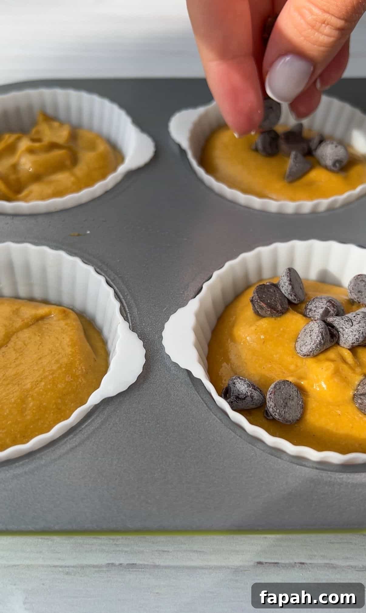 A hand sprinkles chocolate chips over batter in silicone muffin cups placed on a baking tray. The batter is a golden color, filling four cups, ready for baking.