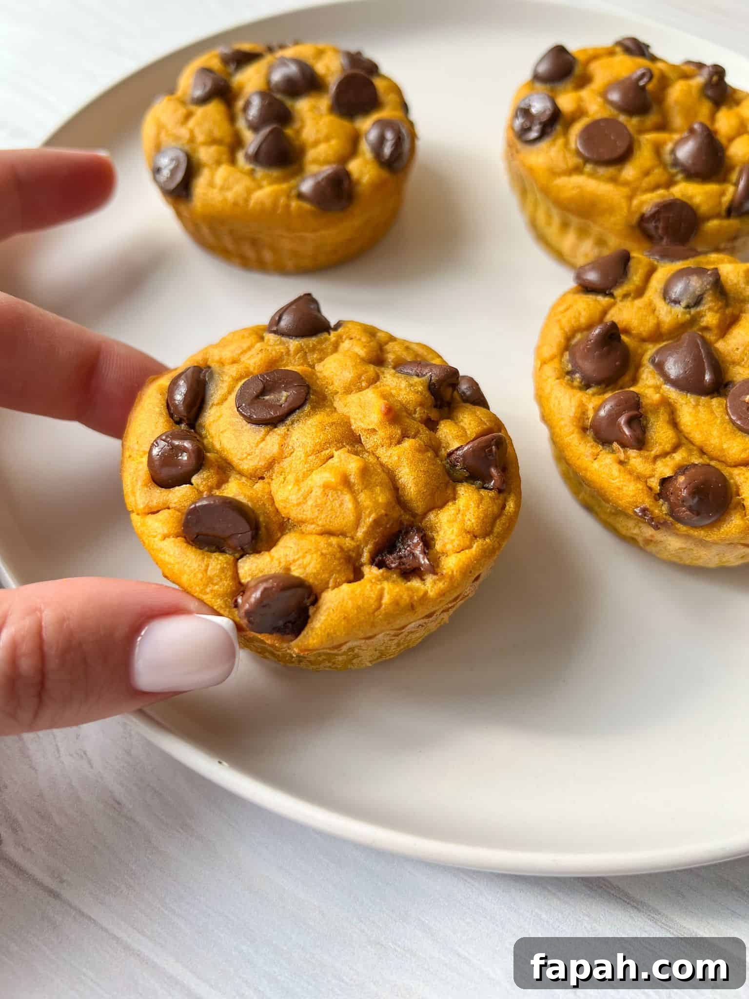 Four chocolate chip muffins are on a white plate. A hand with white nail polish is reaching for one of the muffins. The muffins have a golden-brown color and chocolate chips scattered on top. The background is a light surface.