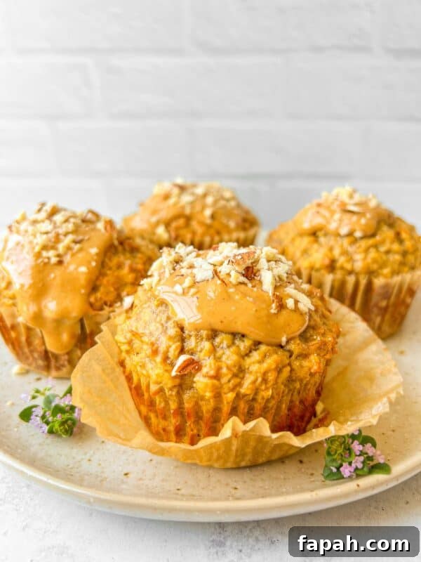 Four muffins on a plate, each topped with a glossy spread and sprinkled with chopped nuts. The plate is garnished with small purple flowers. The background is a white brick wall, adding a rustic touch.