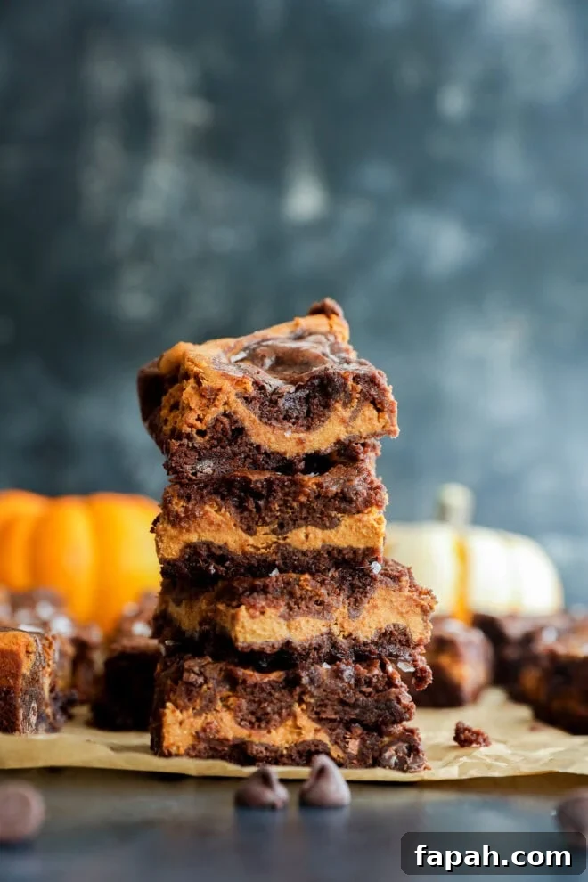 Pumpkin Swirl Brownies with Spiced Cream Cheese Filling 2 A stack of four swirled chocolate and pumpkin bars sits on parchment paper, with chocolate chips scattered nearby and blurred pumpkins in the background.