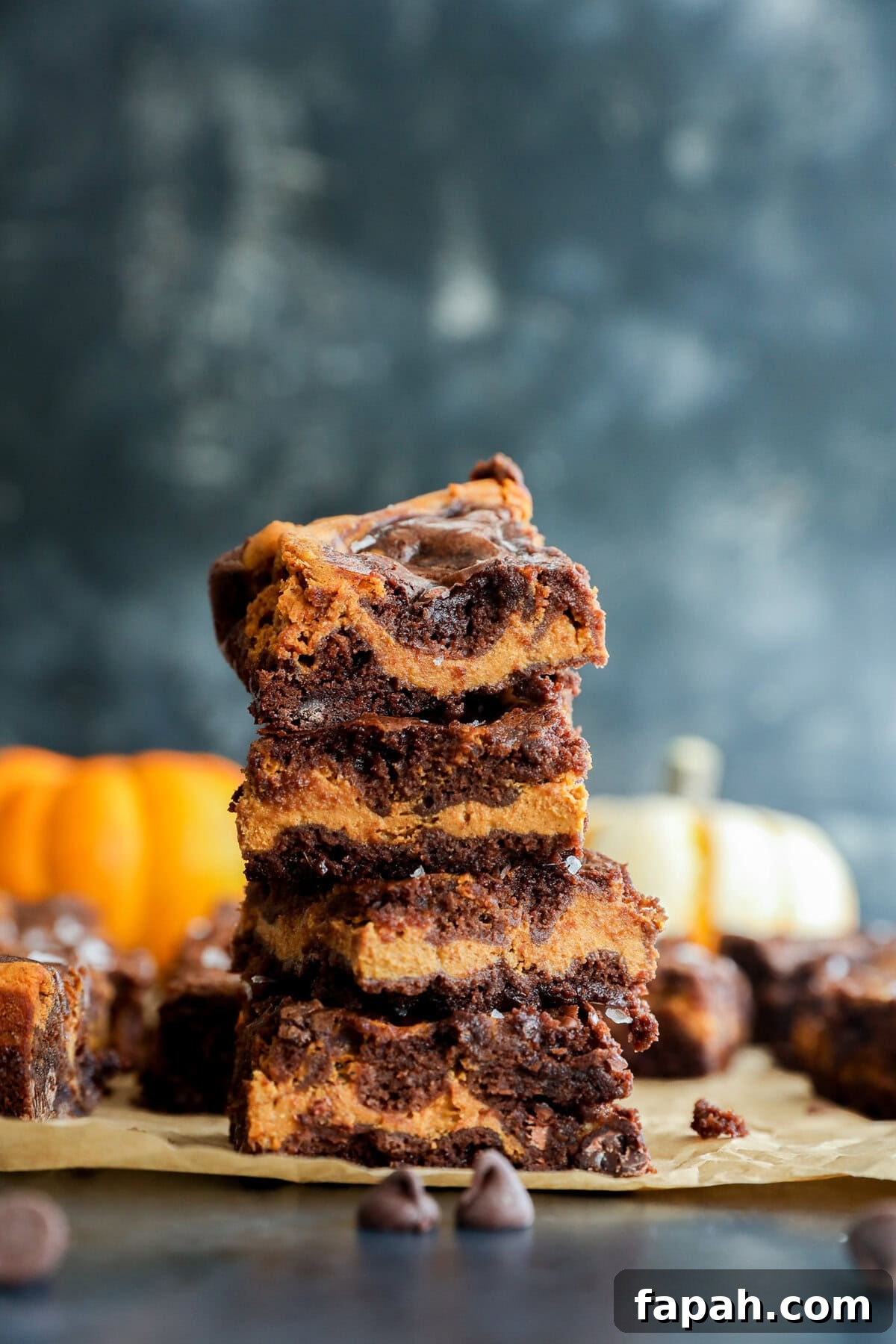 Pumpkin Swirl Brownies with Spiced Cream Cheese Filling 10 A stack of four swirled chocolate and pumpkin bars sits on parchment paper, with chocolate chips scattered nearby and blurred pumpkins in the background.