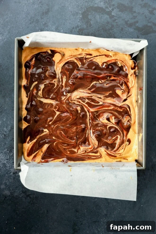 Pumpkin Swirl Brownies with Spiced Cream Cheese Filling 6 A square pan lined with parchment paper holds a freshly baked dessert with a swirled chocolate and light batter pattern on top, resembling brownies or blondies. The background is a dark, textured surface.