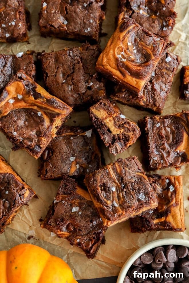 Pumpkin Swirl Brownies with Spiced Cream Cheese Filling 8 A batch of swirled brownies topped with flaky sea salt sits on parchment paper. Some brownies are stacked, revealing fudgy centers. A bowl of chocolate chips and a small pumpkin are in the corner.