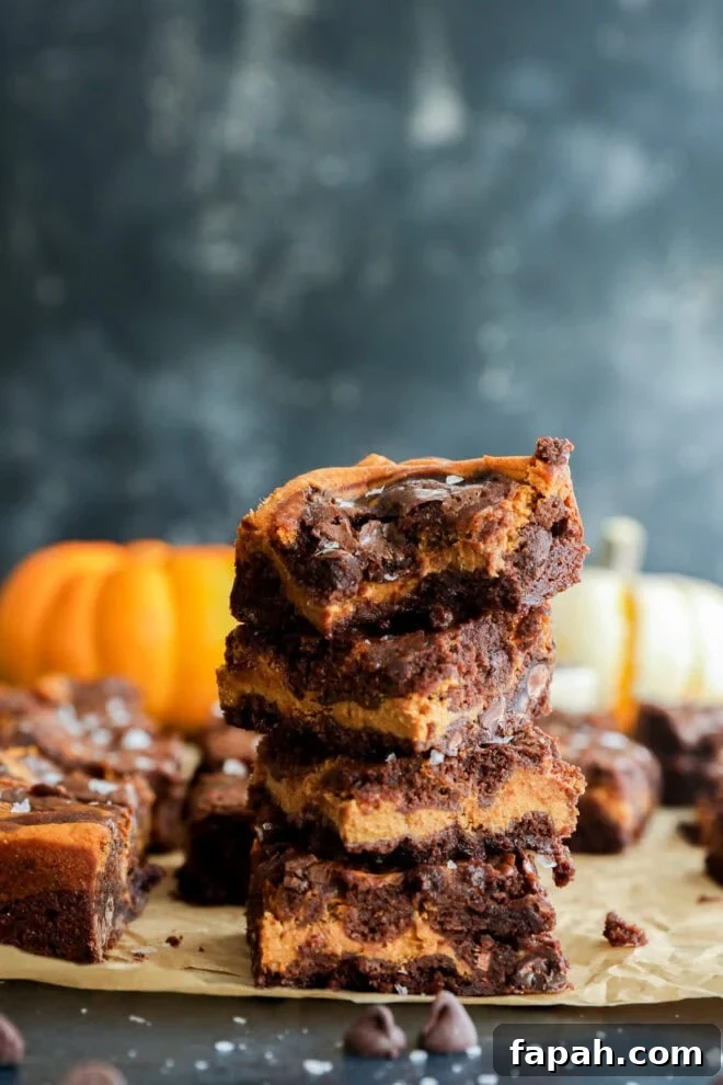Pumpkin Swirl Brownies with Spiced Cream Cheese Filling 9 A stack of four pumpkin chocolate chip brownies sits on parchment paper, with a bite taken out of the top brownie. In the background, there are pumpkins and more brownies, set against a dark, blurred backdrop.
