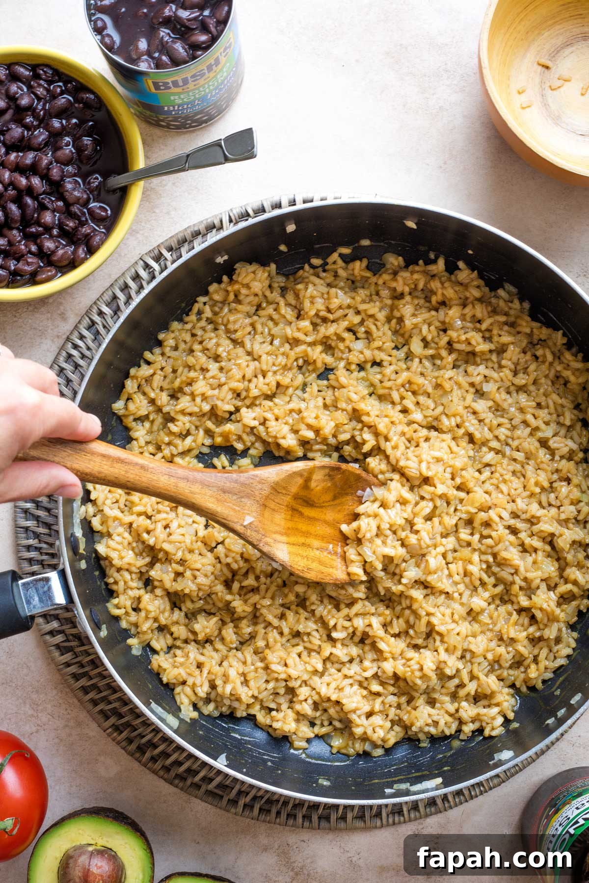 Overhead of step #5 with a hand on wooden spoon, stirring Lizano sauce into the rice in pan.