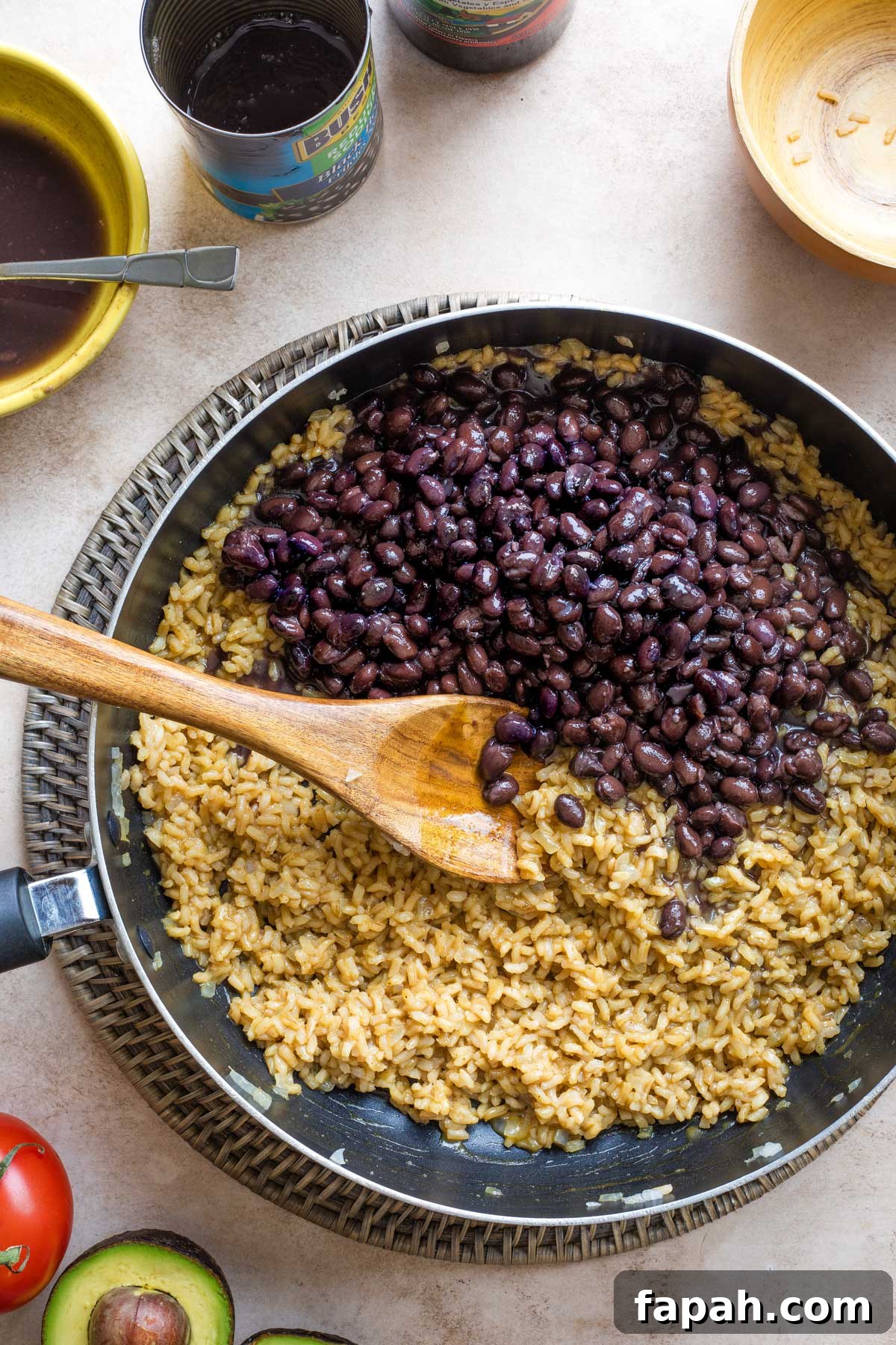 Overhead of recipe step #6 with undrained black beans added to skillet on top of rice; wooden spoon ready to stir them in.