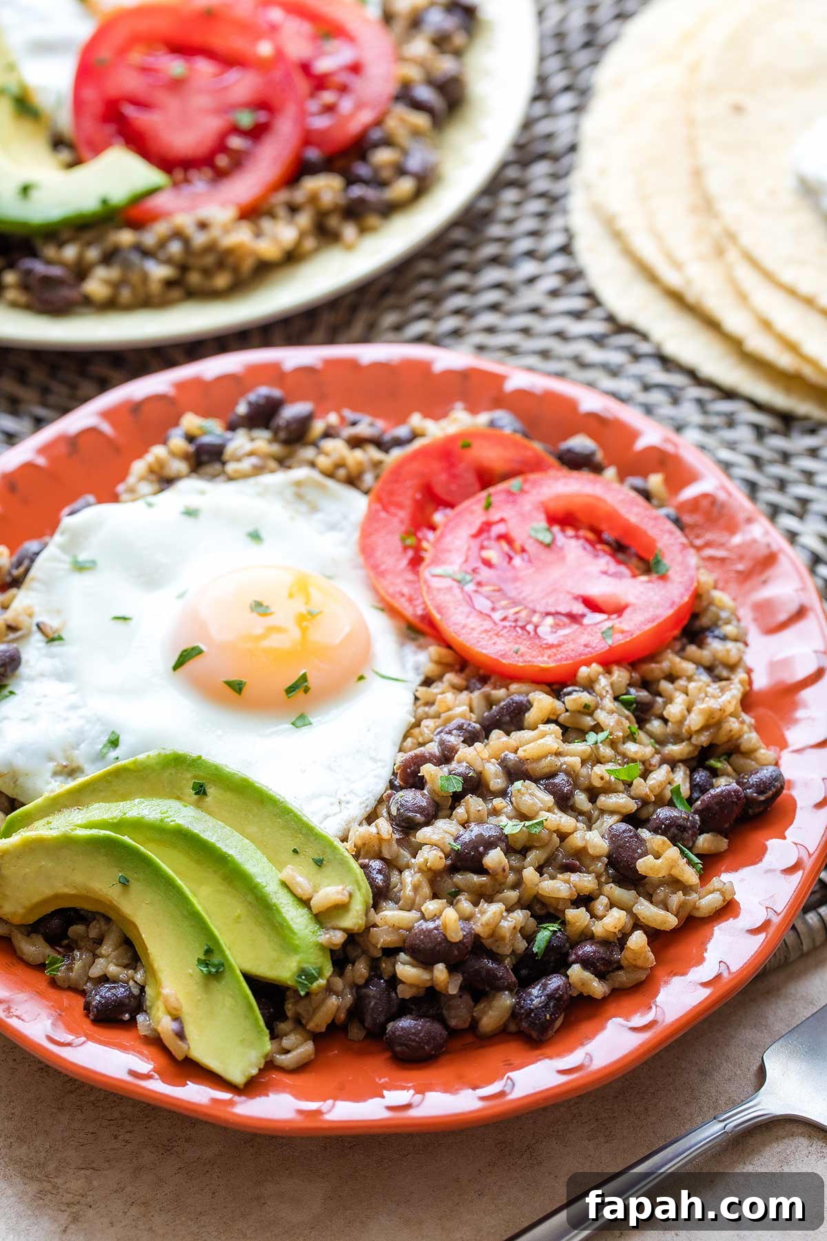 Rice and beans on orange plate with tomato, avocado and an egg nestled on top; second plateful in background.