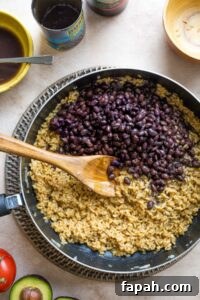 Overhead of recipe step #6 with undrained black beans added to skillet on top of rice; wooden spoon ready to stir them in.