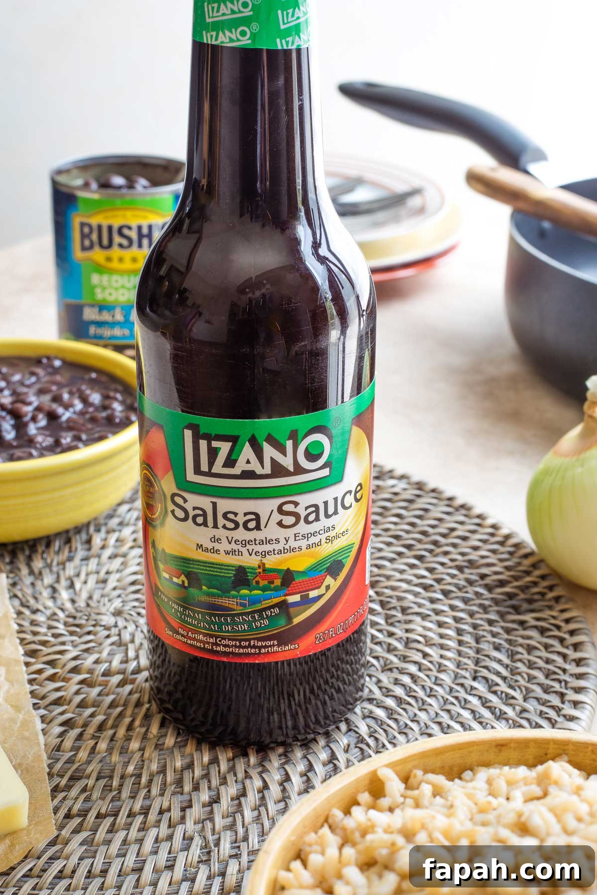 Closeup of a bottle of Lizano sauce with other ingredients like bowls of beans, rice and an uncut onion nearby.