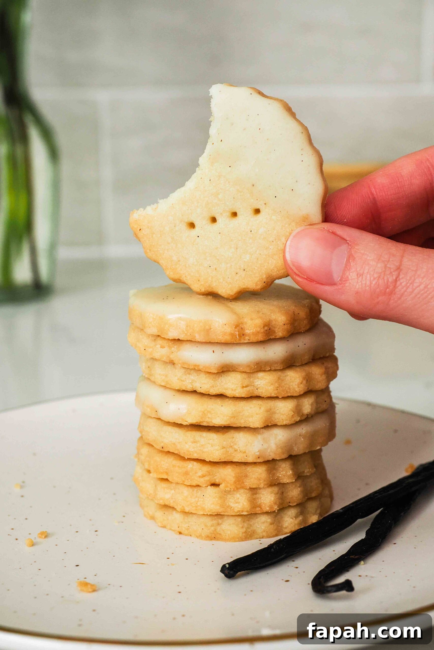 A hand holds up a vanilla shortbread cookie missing a bite in front of a stack of cookies.