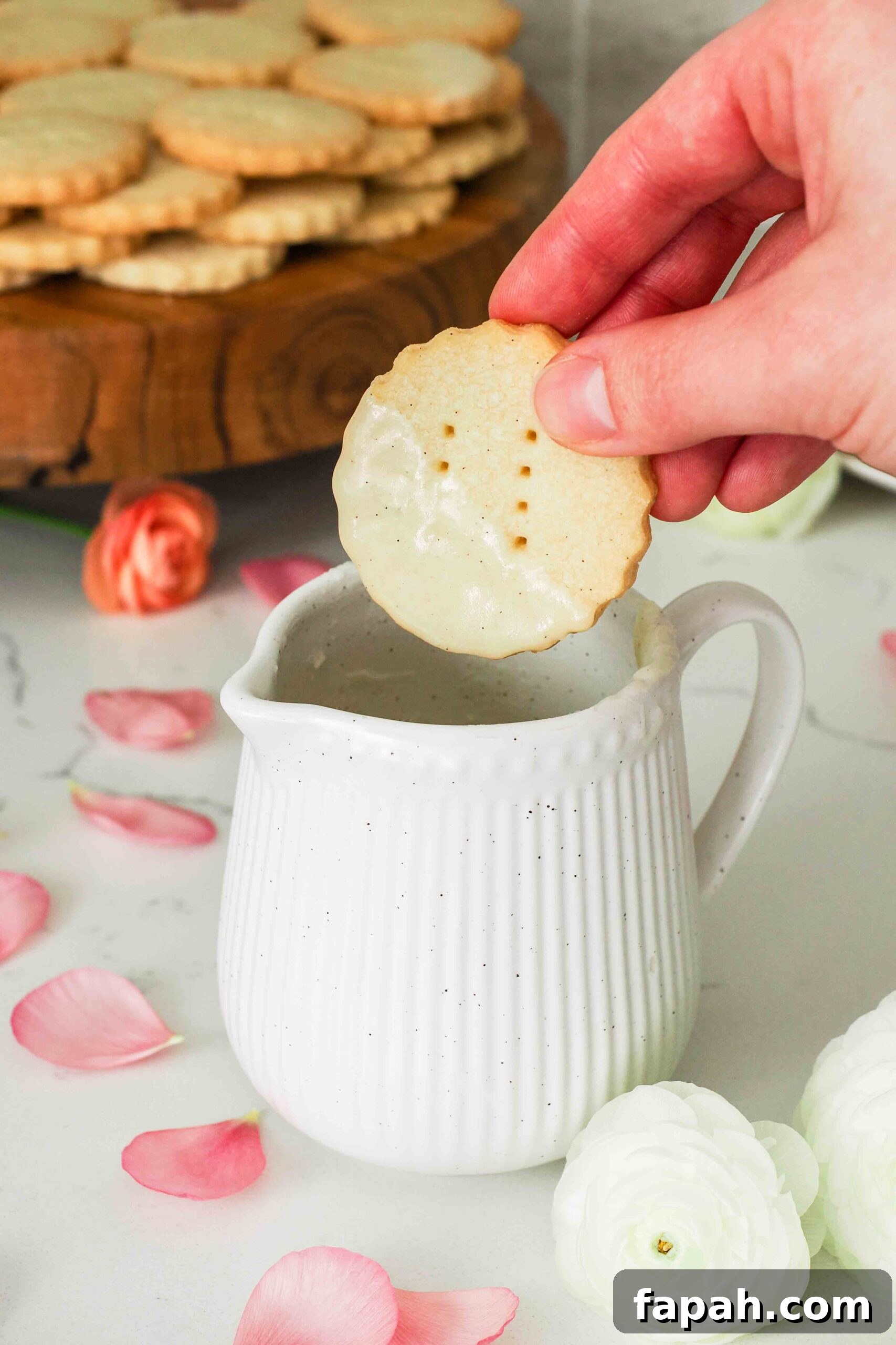 A hand dips a vanilla shortbread cookie into a small jug of vanilla glaze.