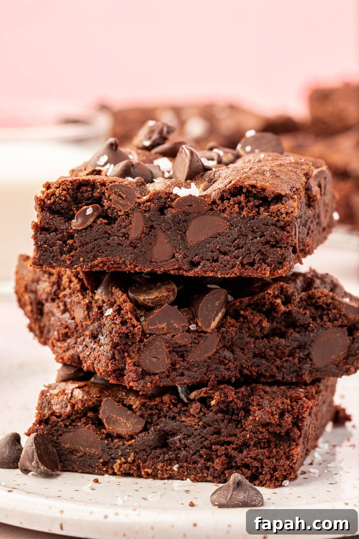 A stack of three gluten-free brownies on a small dessert plate topped with semi-sweet chocolate chips and flaky sea salt.