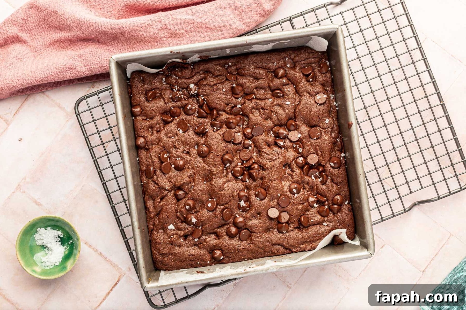 A square baking pan filled with baked gluten-free brownies with a sprinkle of flaky sea salt on top of a pink background.