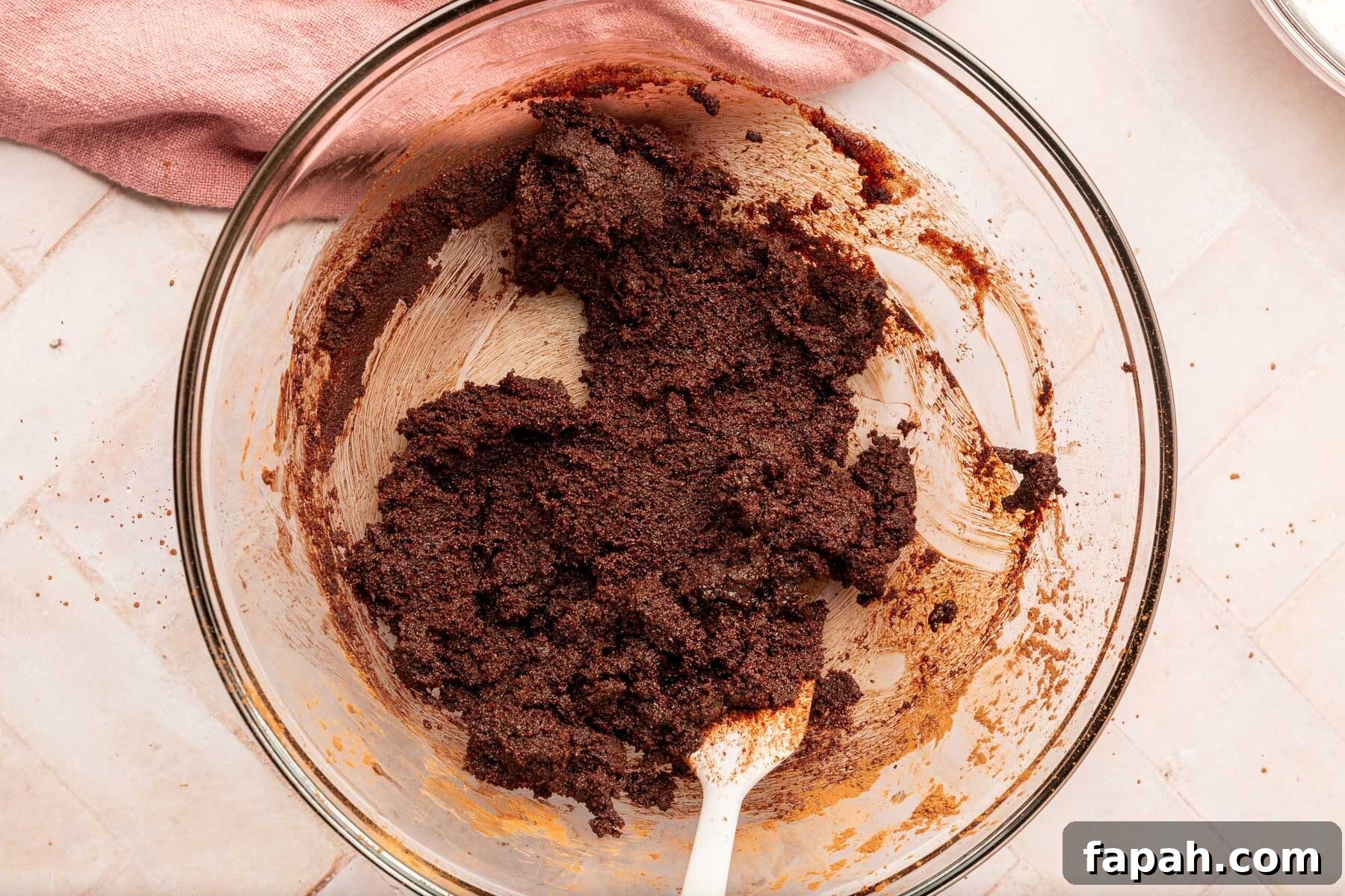 A glass mixing bowl with a chocolate mixture made of cocoa powder, butter and sugar with a pink spatula.