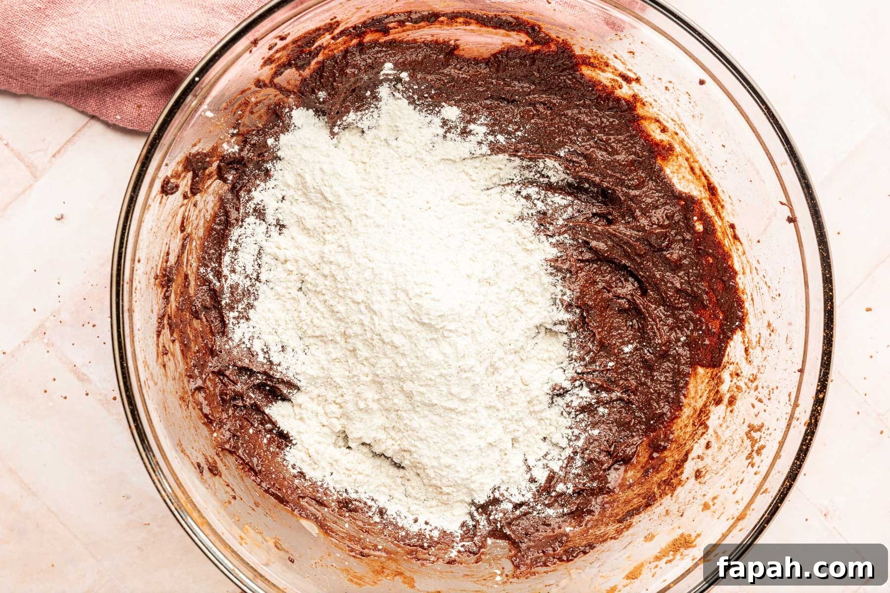 A glass mixing bowl with brownie batter topped with a pile of gluten-free flour before mixing together.