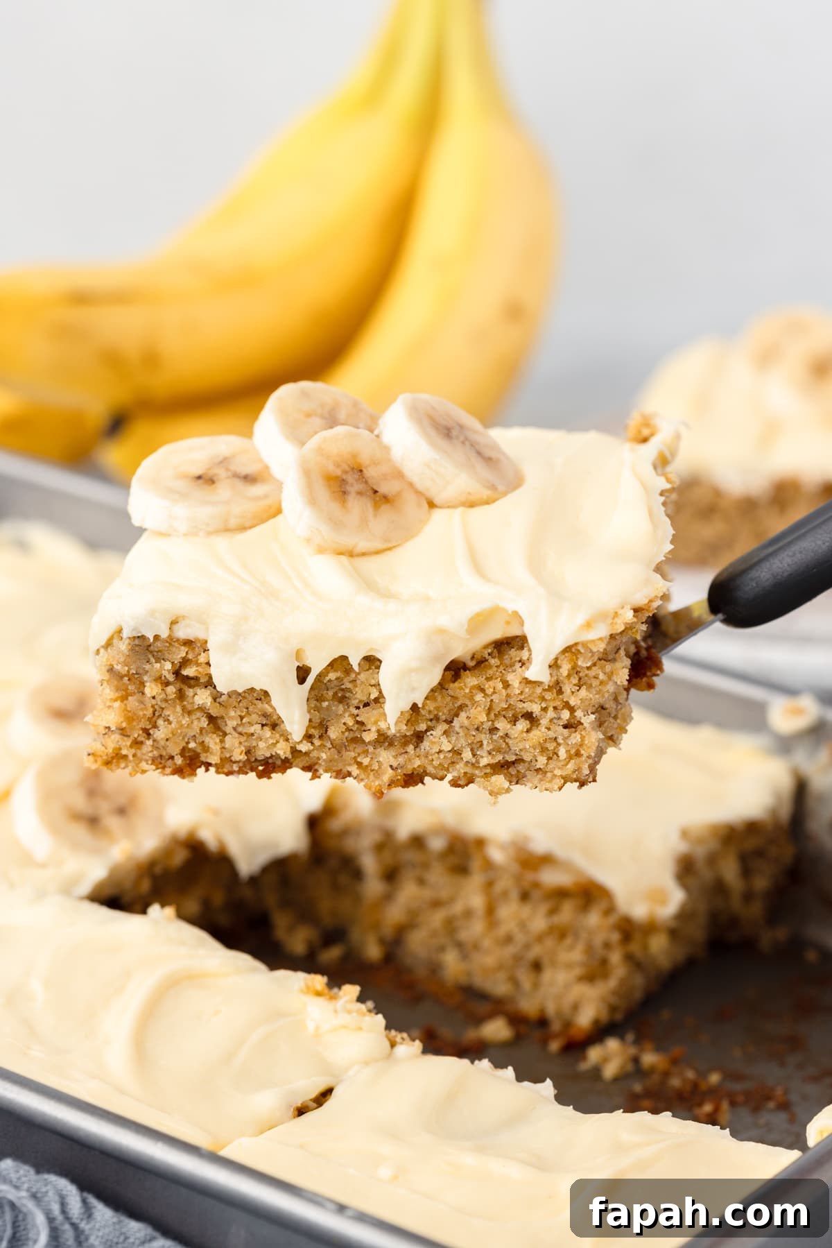 A spatula holds up a slice of fresh milled flour banana cake with cream cheese frosting, topped with banana slices.