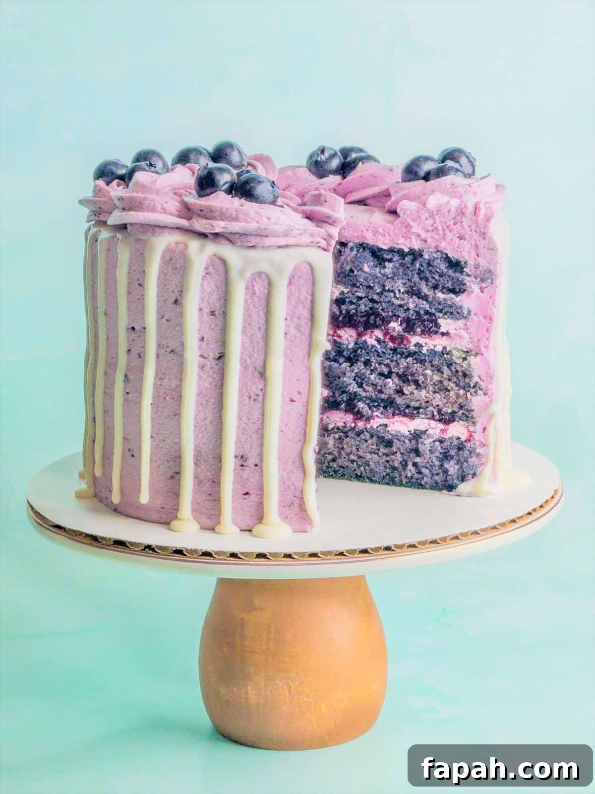 A beautiful blueberry cake on a cake pedestal cut open showing three layers of cake, blueberry filling and blueberry frosting.
