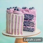 A blueberry cake on a pedestal with a slice taken out showing layers of moist blueberry cake, blueberry buttercream, and blueberry compote filling.