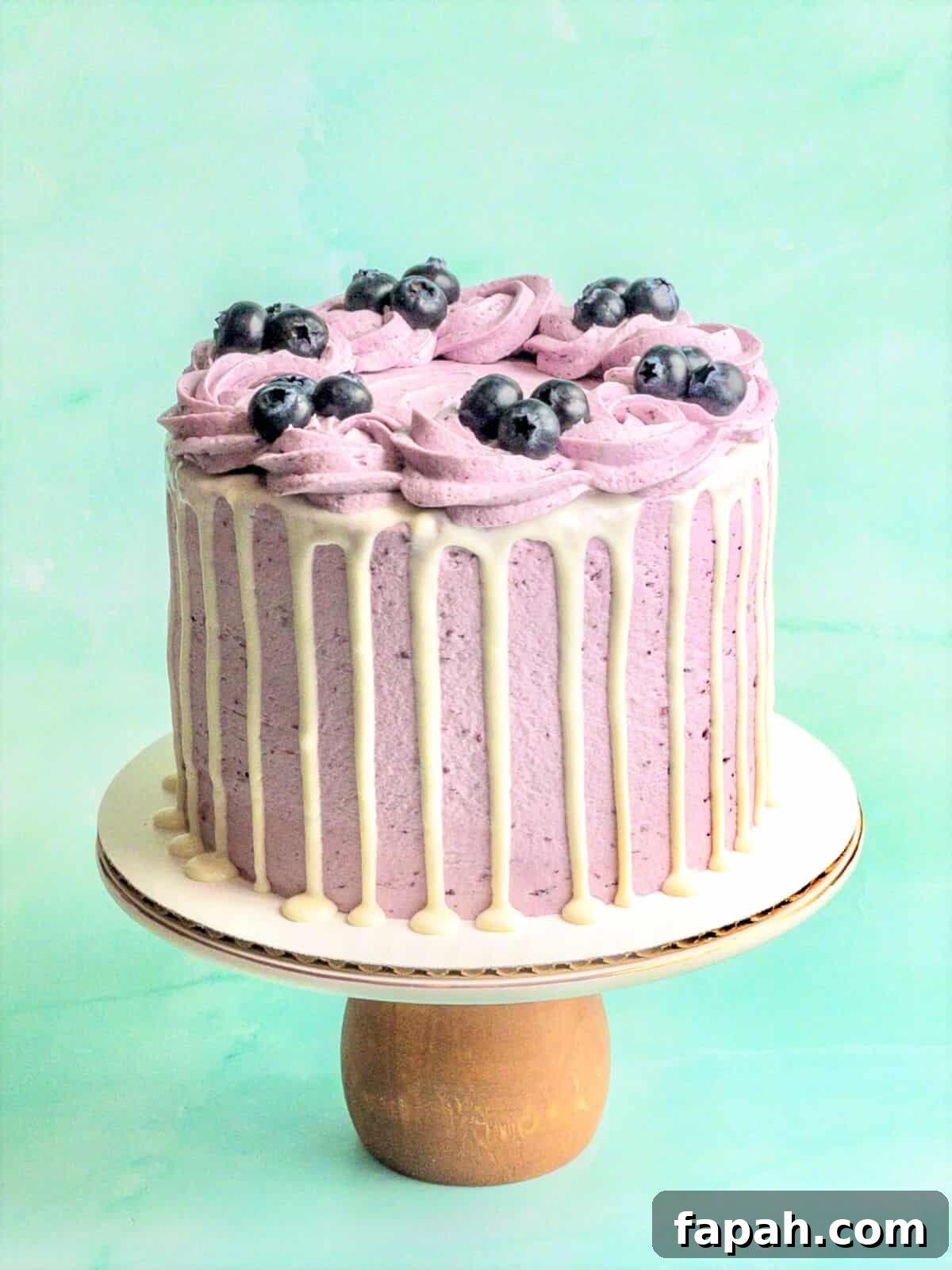A blueberry cake on a cake stand with white chocolate ganache drizzle and fresh blueberries decorating it.