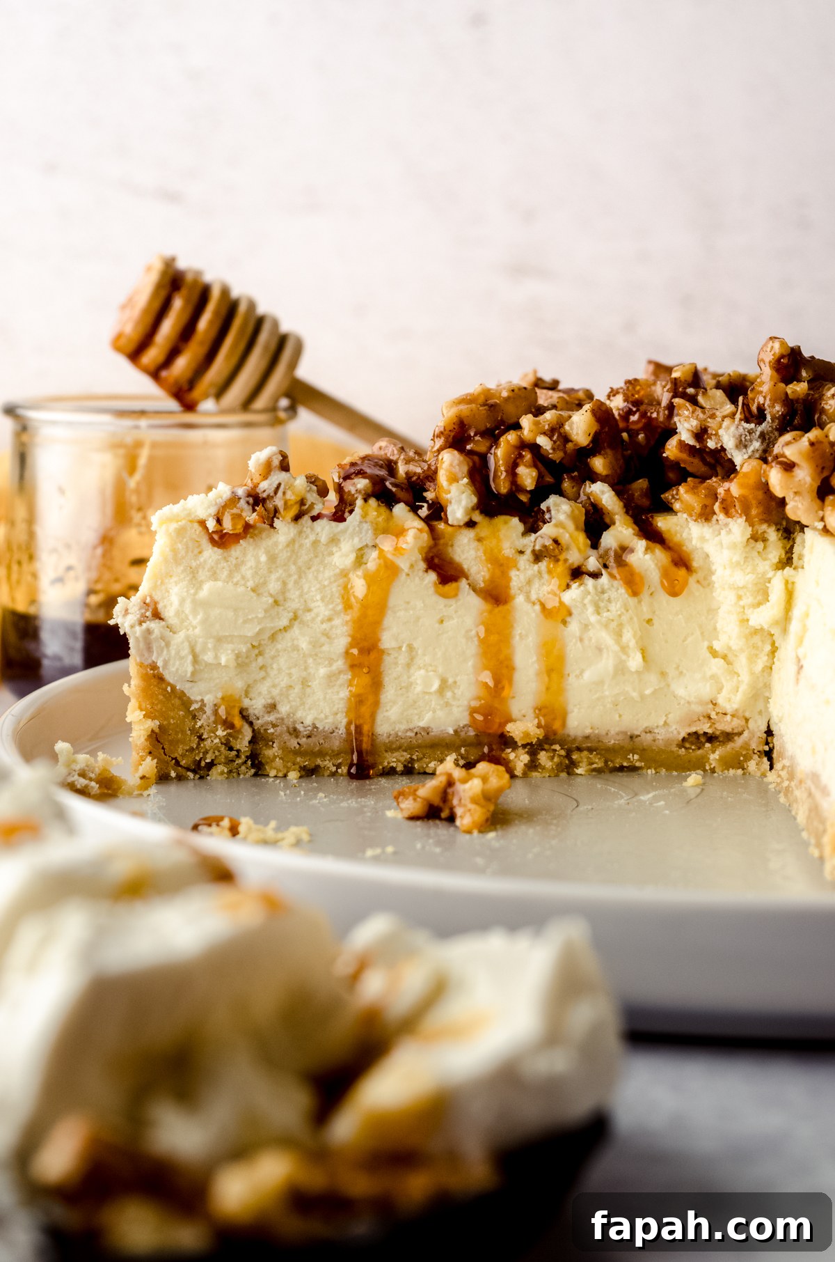 Goat Cheese Cheesecake with Roasted Honey Walnut Topping 2 A sliced goat cheese cheesecake on a plate with a jar of honey and a honey stick in the background and a plate of goat cheese in the foreground.