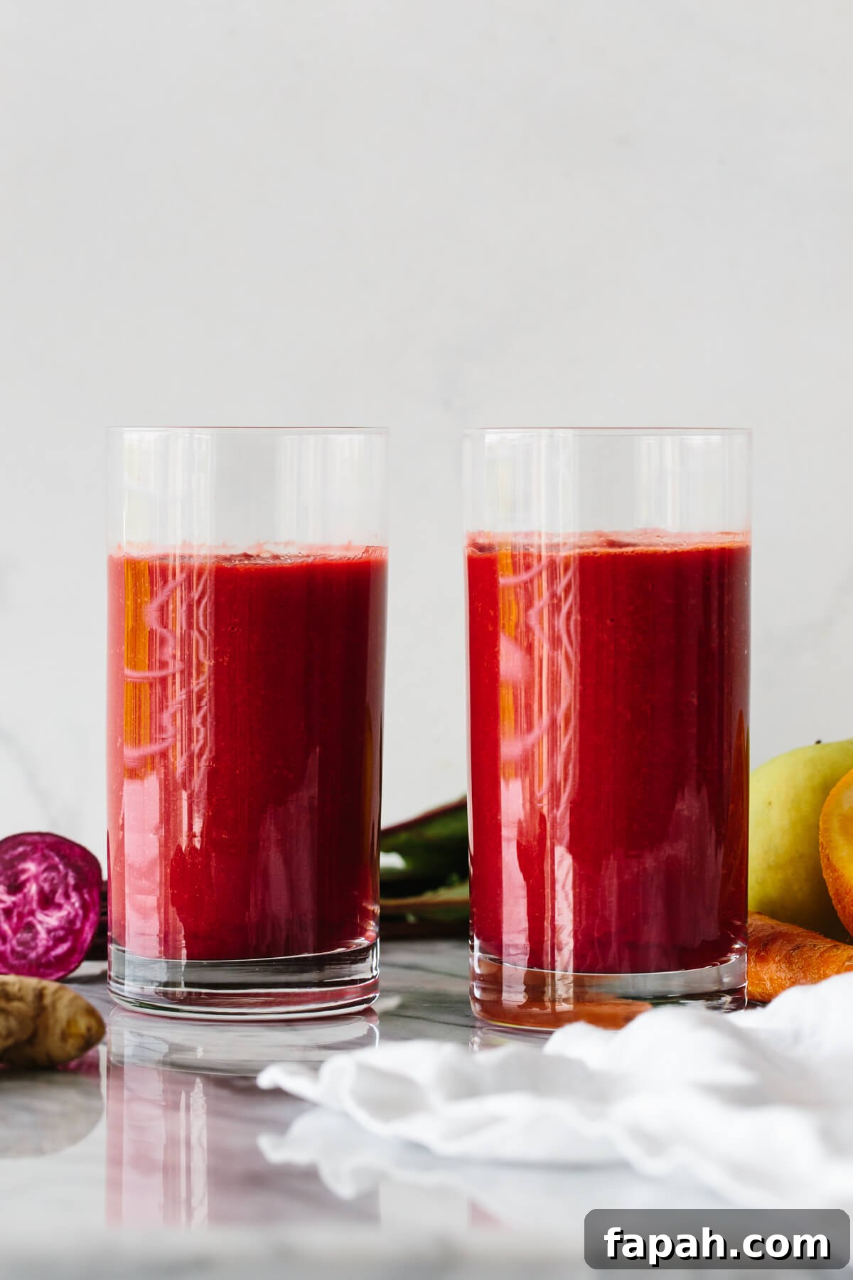 Two glasses of apple, carrot, beet smoothie surrounded by fresh produce.