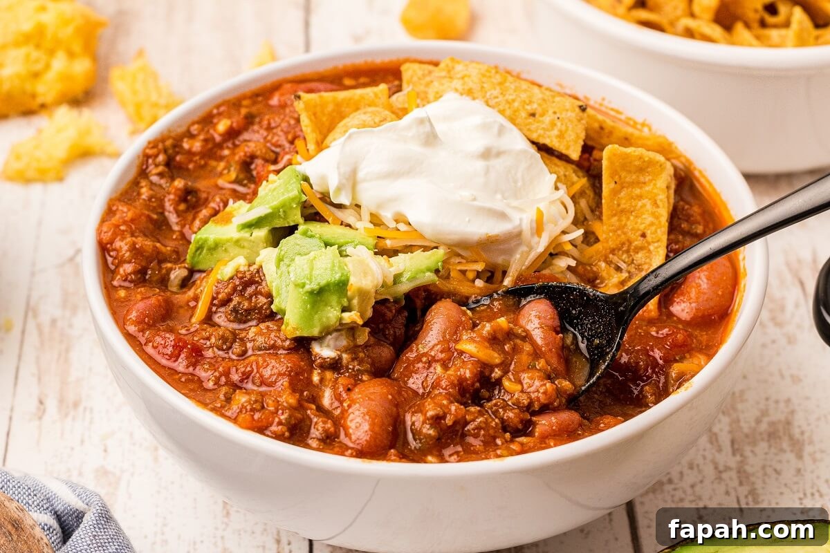 bowl of deer chili with avocado, sour cream, cheese and chips on top. A spoon is digging in.