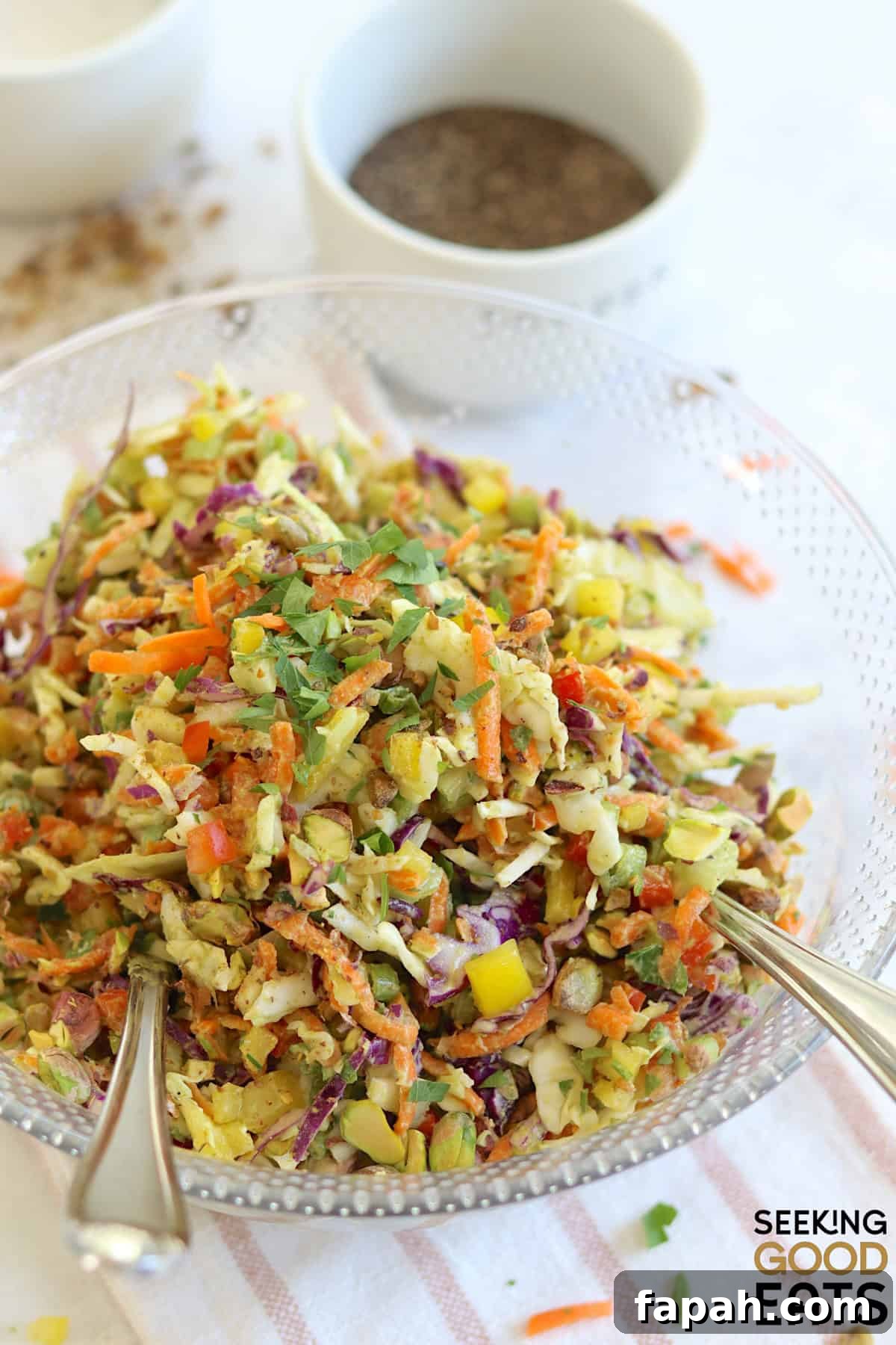 A large round clear bowl filled with green goddess cabbage salad and two silver salad spoons.